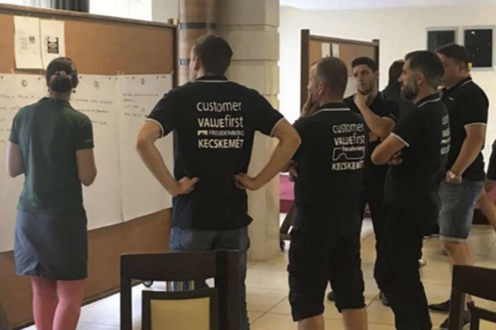 Five men listen to a woman who is explaining something to them on a board.