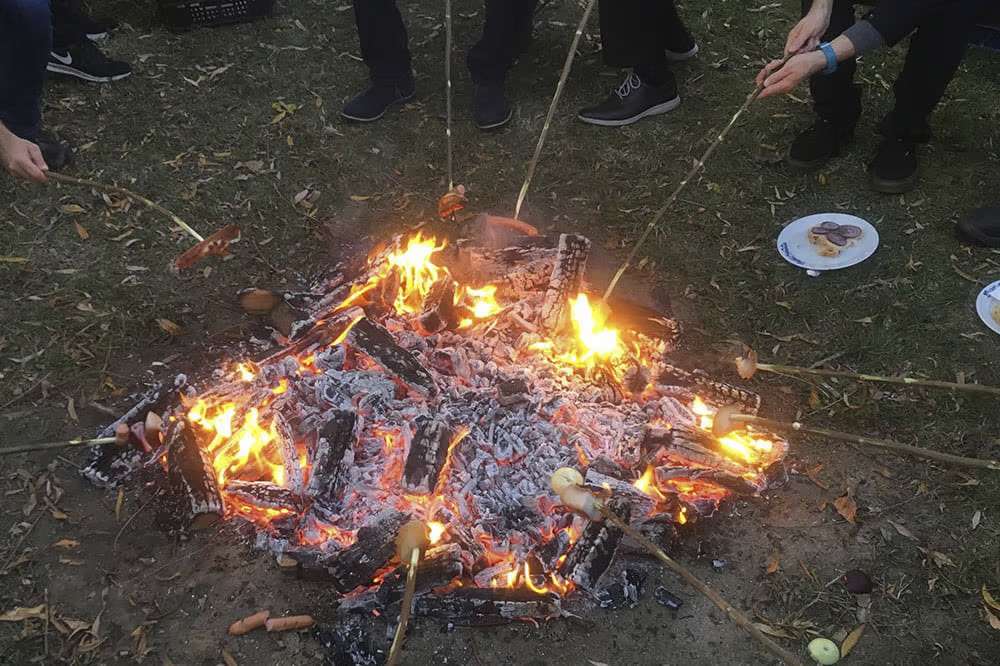 Several hands hold barbecue food on pieces over campfire