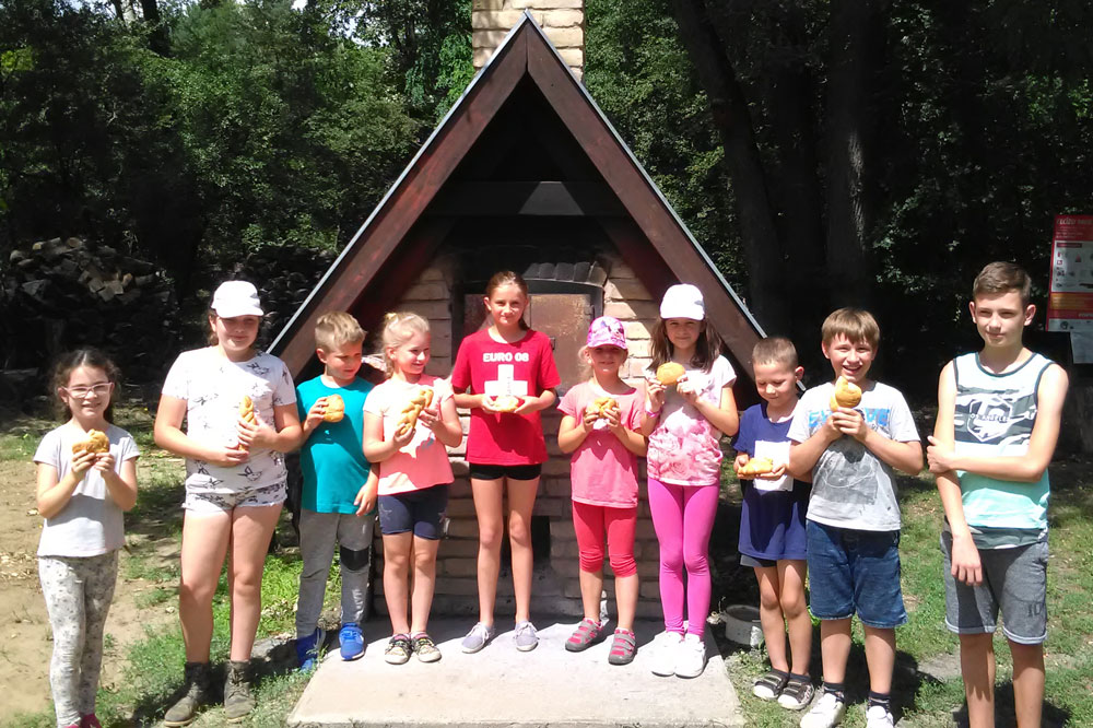 Ten children stand side by side in front of a small triangular house in the park
