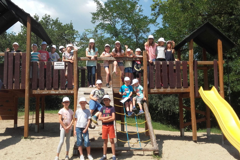 Several children are standing on a wooden castle on the playground and in front of the wooden ramp of the castle there are also a few more children